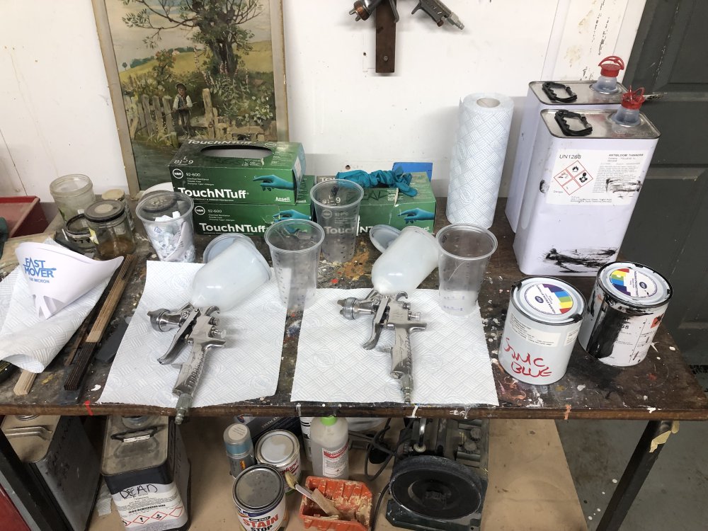 A photo of a paint splattered table on which sit two spray-guns ready to be filled with paint. To the right are two pots of paint, and behind them two canisters of thinner.