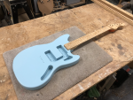 On the workbench sits a work-in-progress guitar, with just the painted-body and neck in position. The body is in the style of a Fender Mustang offset guitar and is painted a pale blue, and the neck is made from birds-eye maple and finished in oil.