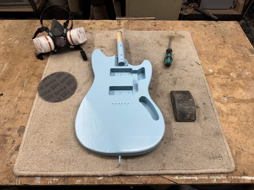 A photo of the offset-style guitar body sat on the workbench, a solid pale blue colour thanks to the painting. In the place where the neck would usually be is a wooden baton that was used to hold it in place whilst it was sprayed. Besides the body sit a screw driver, a sanding block, a sanding disk that says '600' on it, and a respirator mask.
