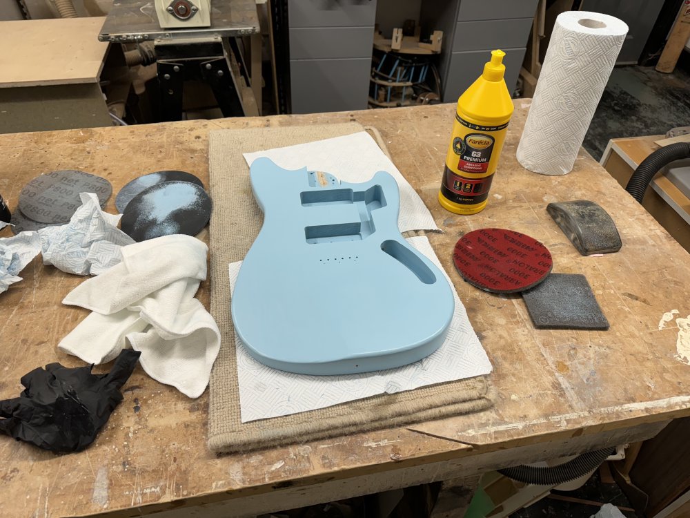A photo of the guitar body on the workbench, still looking a pale blue colour, and it is surrounded by sanding pads that have blue stain on them, and a bottle of the abrasive compound can be seen to one side.