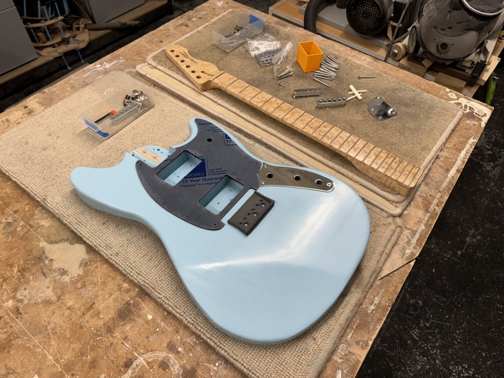 The pale blue guitar body sits on the workbench, and on it sit a black pick-guard (still in protective plastic wrap) and the chrome plate control cover. Next to it sits the birds-eye maple neck, and then a whole pile of screws and parts such as the neck plate, the nut, and the string-ferrule-block.