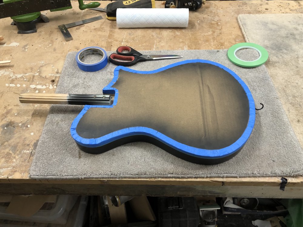 A photo of a guitar body on the workbench with a cardboard cover stuck on the front with masking tape.