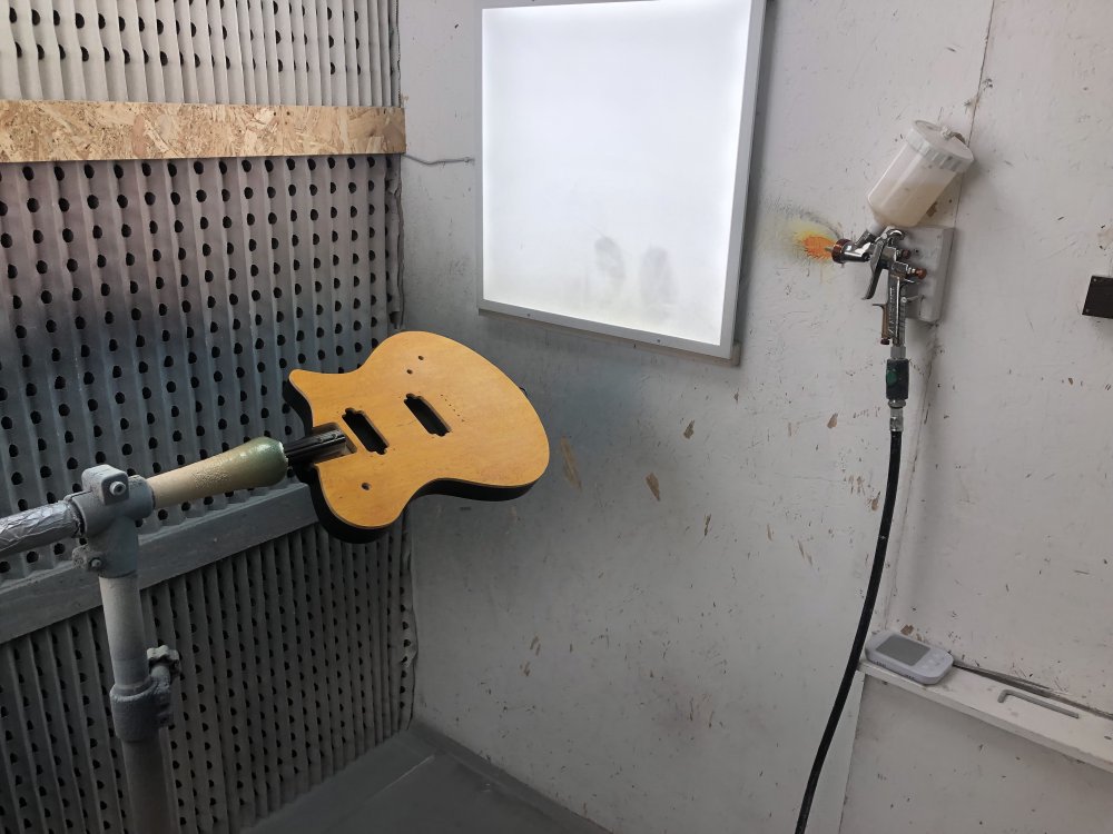 A photo of the guitar body with the yellow front and black rear mounted in a stand in spray-booth. On the wall is a spray-gun with a half-filled pot of clear lacquer on it.
