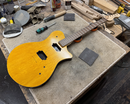 A photo of a work-in-progress guitar on the workbench, surrounded by tools and sanding pads. The guitar has a maple neck with a rosewood fretboard and 21 frets, and the body is a bright yellow stain on birds-eye maple on the front, with black and natural wood sides. There are holes in the body for components, but all are missing.