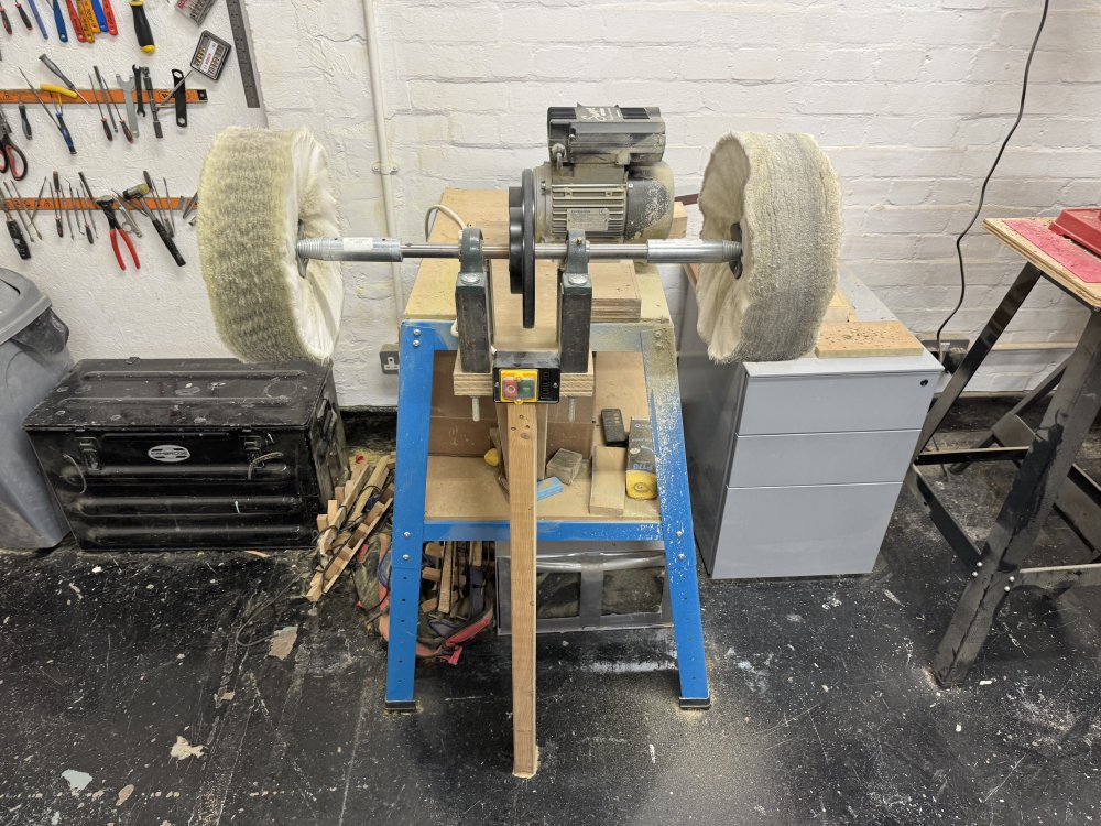 A photo of a buffing machine, which has a bar mounted at about waist height off the floor, attached to a motor by a belt. On either end of the bar are large wheels of soft fabric.