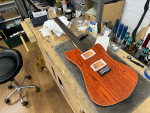 A mostly assembled guitar sits on the workbench! It is missing all the electronics, but otherwise looks like a guitar. Importantly it has strings on it! It has a vivid orange-stained wood body with white binding and white pickup mounts, and a maple neck with a rosewood fretboard.