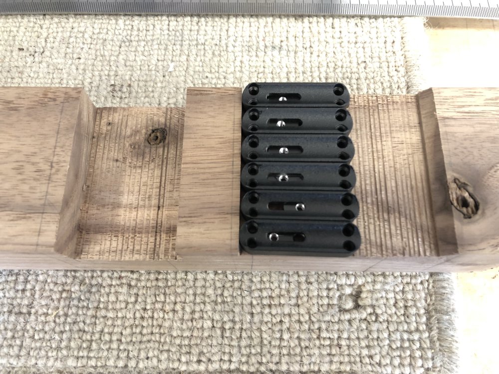 A photo of six black blocks mounted on some wood that will later become a guitar. The black blocks are lozenge shaped if viewed top down, have four holes in the corners, and a slot down the middle. In the slot you can see another bit of black metal with a screw hole in it.