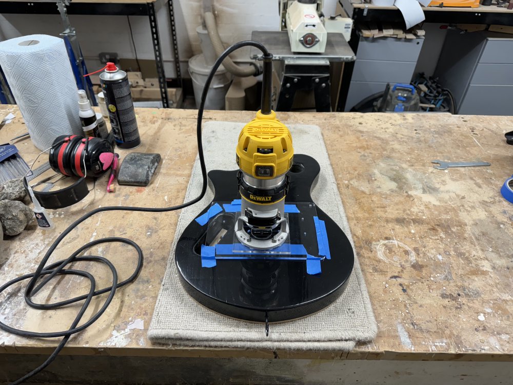 A photo of a painted and varnished guitar body sat face down on the workbench with a palm-router sat on it. Between the two are some clear acrylic templates held down by masking tape.