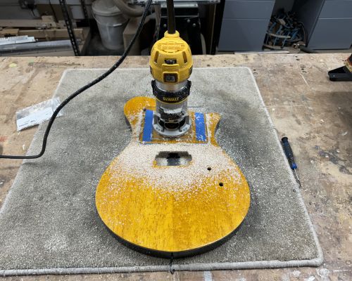 A photo of a yellow and black finished guitar body on a workbench, on top of which is a palm-router and everything is covered in wood-dust from some cutting.