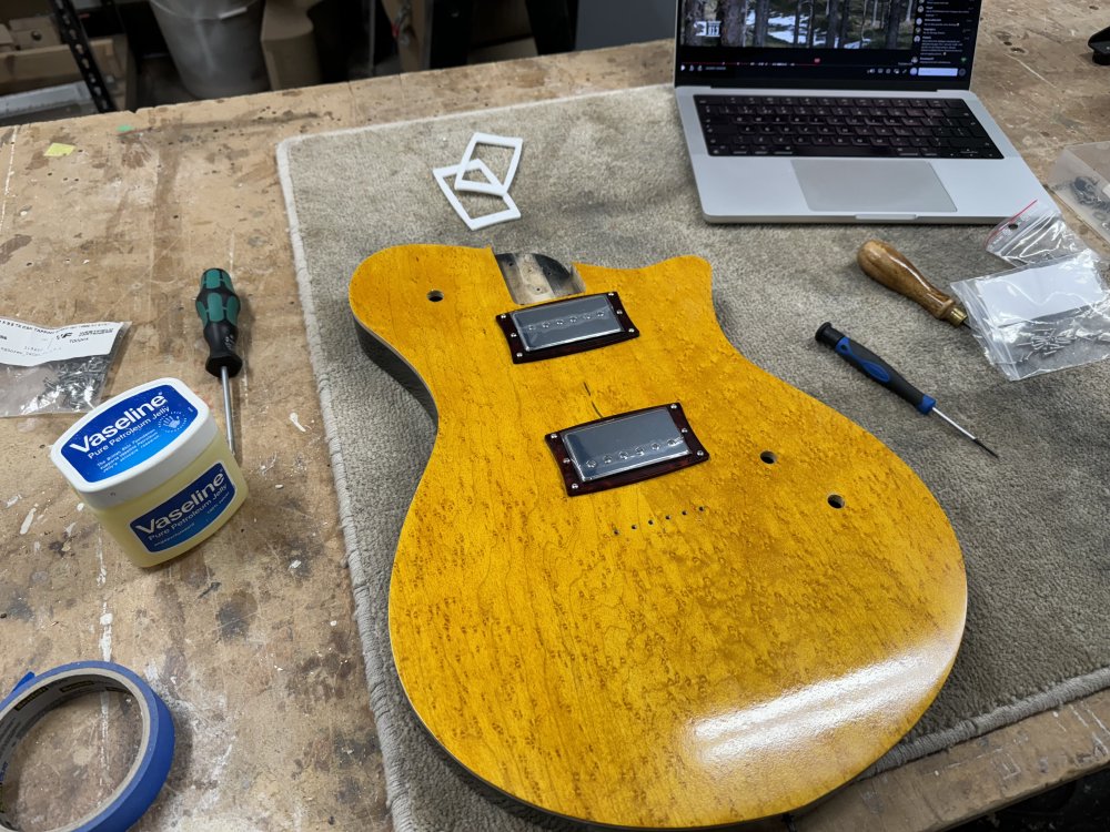 A photo of a yellow and black finished guitar body with some chrome covered humbucker-sized pickups in place with their pickup mounting rings. To the side is a pot of vaseline, looking slightly out of place in the workshop.