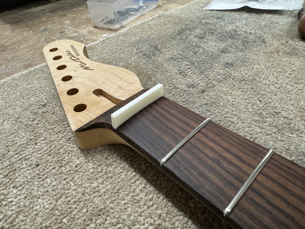 A photo of the top end of a guitar neck, showing an rectangular, oversized nut-blank installed where the nut should be.