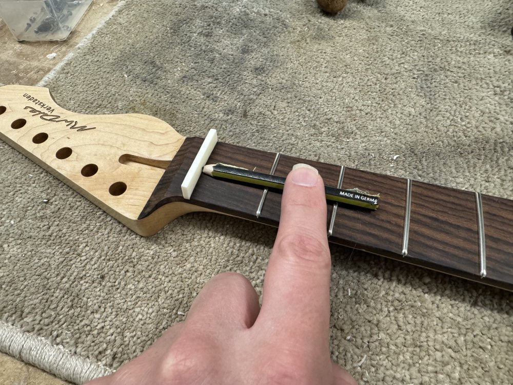 A photo of the neck and oversized nut blank again, but now suspended across the top two frets is a pencil that has been cut in flat length ways, so that the tip is level with the top of the frets. It is being used to draw a line on the nut that matches the height of the frets.
