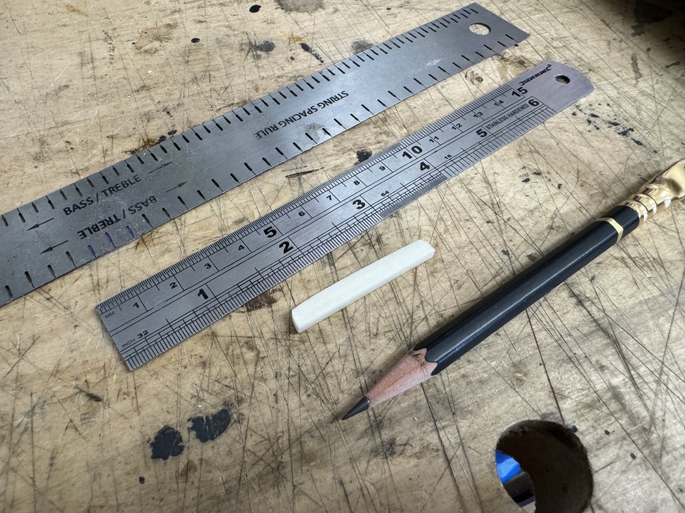 A photo of the nut-blank, now cut to the right height, on the workbench next to a regular pencil, a regular ruler, and a string spacing ruler that has notches in it where you can draw through the ruler at string spacing intervals.