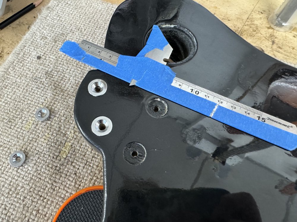 A photo of the back of a guitar body near the neck pocket, and on that is a ruler to which has been taped a razor blade so that only a tiny bit of the blade protrudes. It is next to a series of recessed circles where the neck bolts will go through.