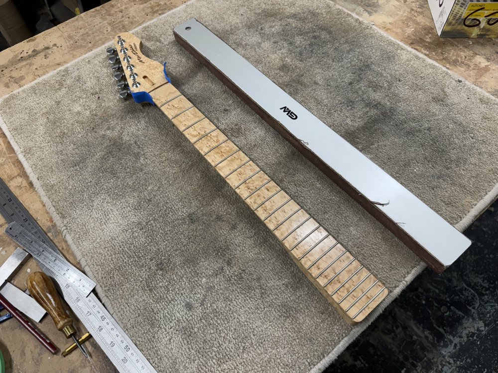 A finished birds-eye maple neck sits on the workbench, and next to it is a long straight metal bar with sandpaper stuck to one edge.