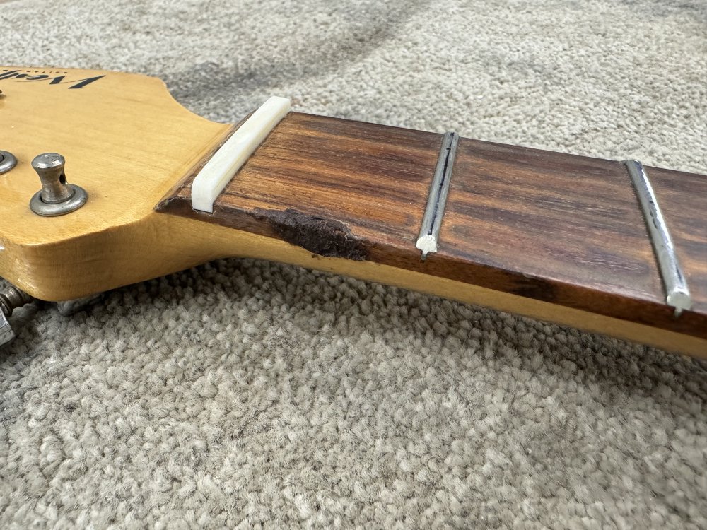 A photo of the top end of a maple neck with a rosewood fretboard. On the side of the fretboard between the nut and first fret is a brown mess where some goop has been applied and hardened.