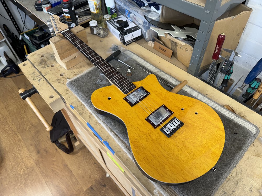 A photo of a solid-bodied electric guitar on the workbench, looking nearly complete, just with holes where the pickup selector switch and the volume and tone controls should be. The neck has a rosewood fretboard on a maple body, and the guitar body is a bright yellow-stained wood finish with black sides. Next to the guitar on the workbench there are two T shaped hole reaming tools.