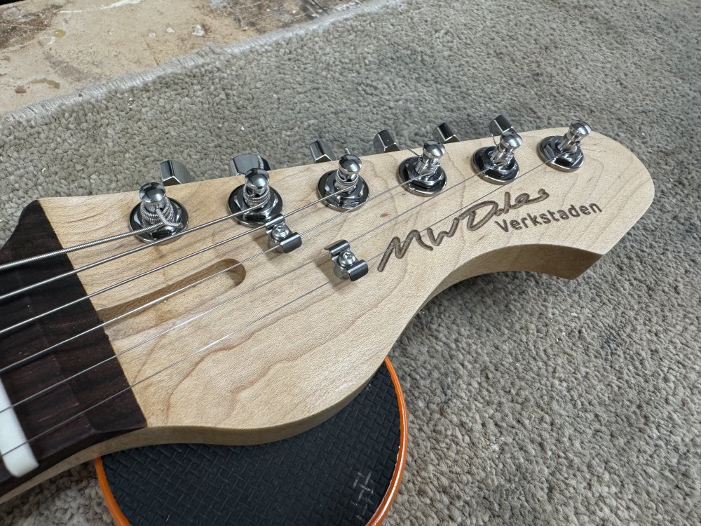 A close-up photo of the headstock of the guitar, which shows the maple neck wood, the tuners, and the two string-trees. Laser-etched into the headstock is 'M W Dales' in handwritten script, and 'Verkstaden' in a san-serif font.