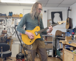 A photo of me stood in the workshop playing a solid-bodied electric guitar that has a bright yellow wood stained body, a rosewood fretboard and a maple neck. I appear to be playing an A barre chord.