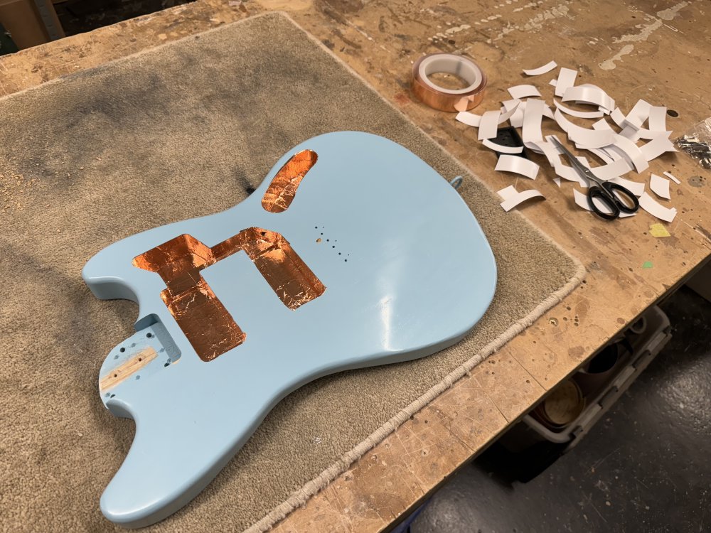 A photo of an offset-style solid-body electric guitar body on the workbench. The body is painted light blue and inside all the cavities cut out for parts to be added it is lined with copper tape that is reflecting the light to create a glow. Next to the body is a lot of bits of tape backing cast to the side.
