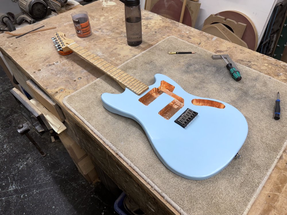 A photo of the guitar body now with a birds-eye maple neck attached, which has tuners, frets and a nut installed. The bridge plate now has chrome plated saddles installed too.
