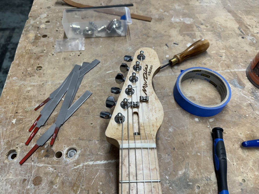 A close up photo of the headstock of the guitar, showing the brids-eye maple in detail. You can see the tuners, the nut and the two strings trees (along with the strings). Laser etched into the headstock is 'M W Dales' and 'Hästen'. Next to the headstock on the workbench are a set of tiny files for cutting slots into the nut.