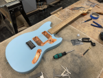 A photo of a work-in-progress solid-body electric guitar laid on a workbench in a workshop. The guitar has a body painted light blue with just a bridge installed and no electronics or pick-guard yet, showing the internal cavities lined with copper tape. The neck is made from birds-eye maple. Most importantly the guitar has strings on it.