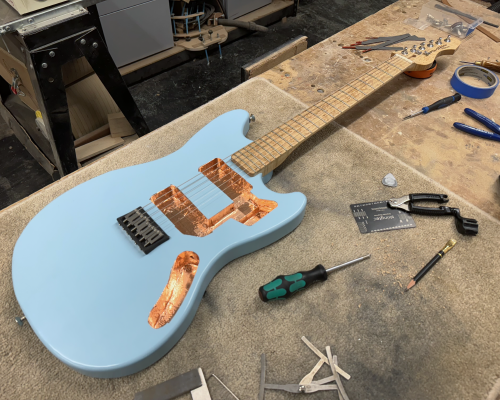 A photo of a work-in-progress solid-body electric guitar laid on a workbench in a workshop. The guitar has a body painted light blue with just a bridge installed and no electronics or pick-guard yet, showing the internal cavities lined with copper tape. The neck is made from birds-eye maple. Most importantly the guitar has strings on it.