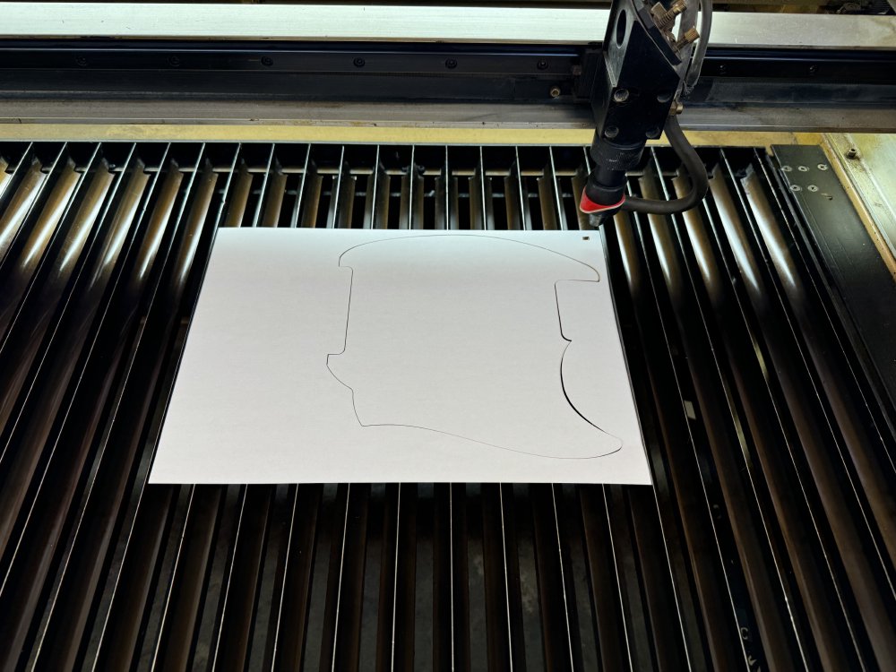 A photo of a sheet of A4 paper on the bed of a laser-cutter, with the outline of a guitar pick-guard marked into it.