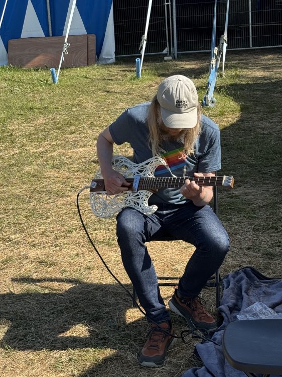 A photo of me sat on a chair on some grass next to a circus-style tent, playing the same unusual looking guitar from the previous photo.