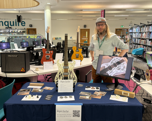 A photo of me stood in a library behind a table with several guitars, including one where the body is 3D printed.