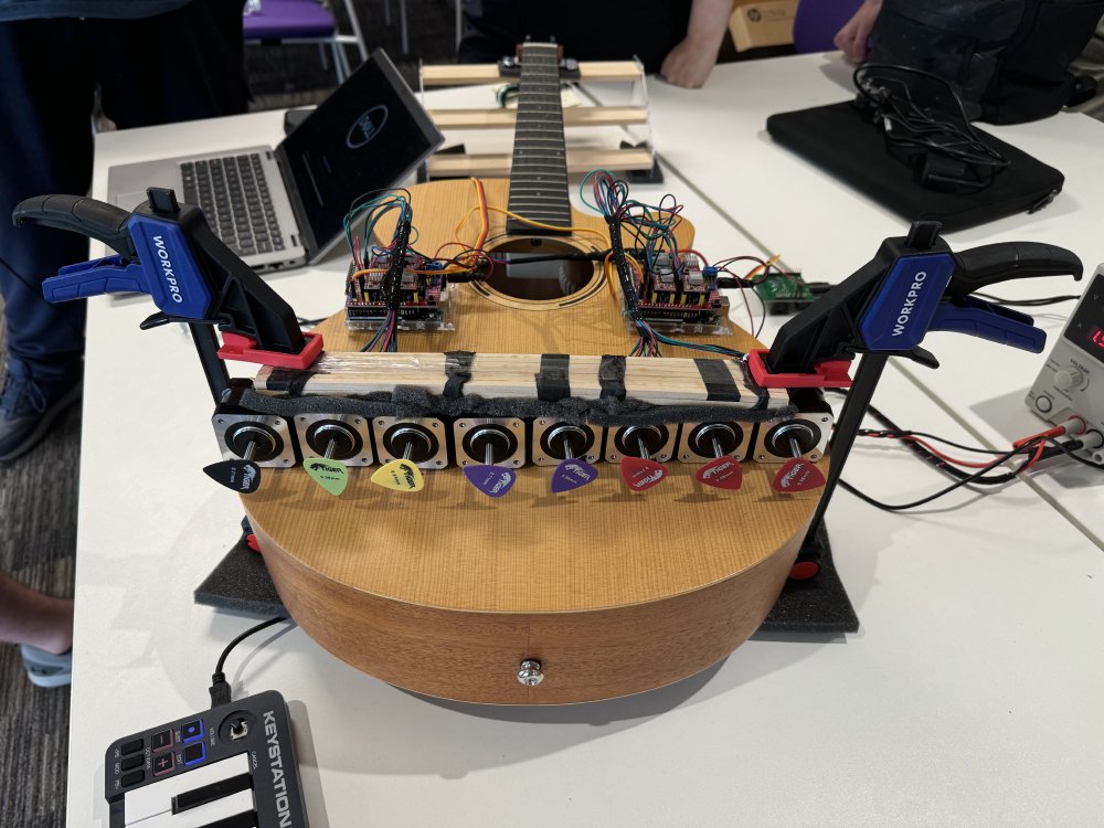 A photo of an acoustic guitar without strings laying on a table. Mounted to the front of the guitar using a pair of G clamps is a strip of eight stepper motors, each with a different coloured plectrum stuck to the end. Also on the guitar are a pair of raspberry pi sized development boards connected to the motors, and wires can be seen going from the boards to a laptop.