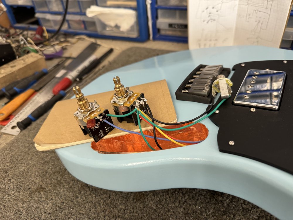 A close up photo of the volume and tone pots being soldered to the wires. Because the wires are running through the body now and quite short, the pots are sat on one of the notebooks next to the cavity in which they'll be mounted (the notebook keeping the components and any solder splash off the body's paint).