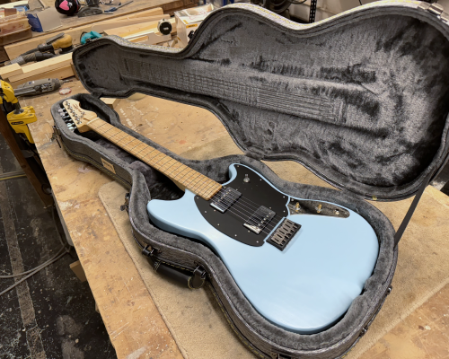 A photo of an electric guitar sat in a case on a workbench in a workshop. The guitar is an offset design, with a pale blue painted body, a black pickguard, with two chrome humbucker pickups on it. The neck is a birds-eye maple.