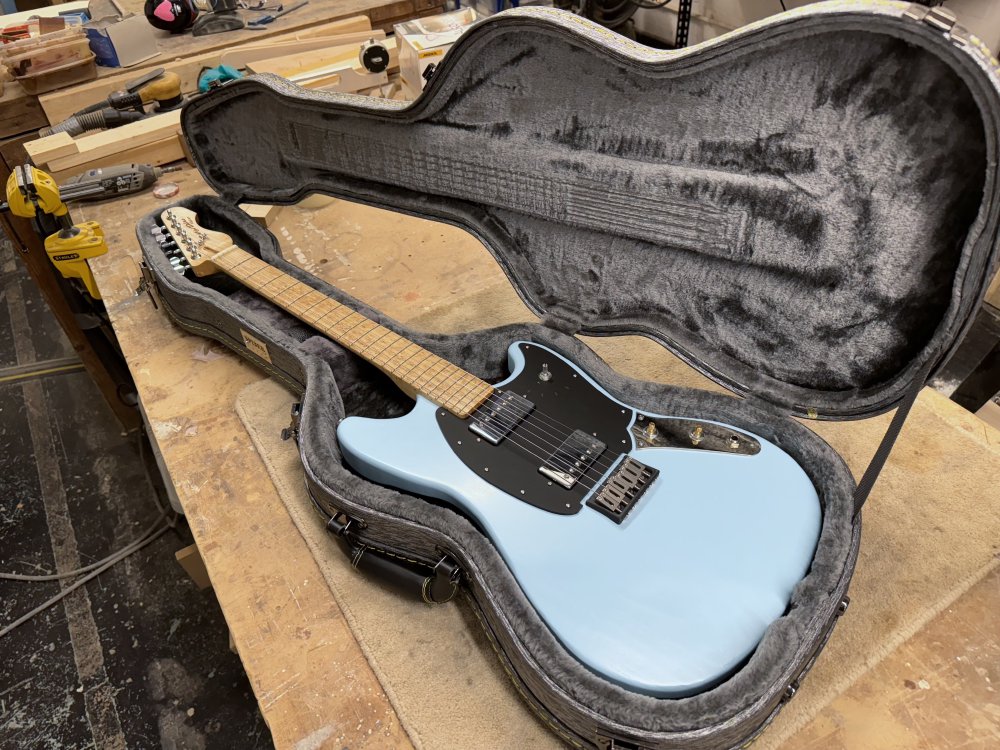 A photo of an electric guitar sat in a case on a workbench in a workshop. The guitar is an offset design, with a pale blue painted body, a black pick-guard, with two chrome humbucker pickups on it. The neck is a birds-eye maple.