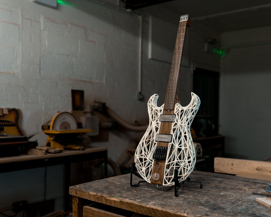 A photo of an unusual looking guitar siting on a stand on a workbench in a moodily lit workshop. The neck and the centre of the body along the neck are made from a single piece of dark walnut wood, whilst the sides of the body are made from a white 3D-printed lattice work. There is no headstock, just a custom aluminium block to anchor the strings, with tuners being built into the bridge.