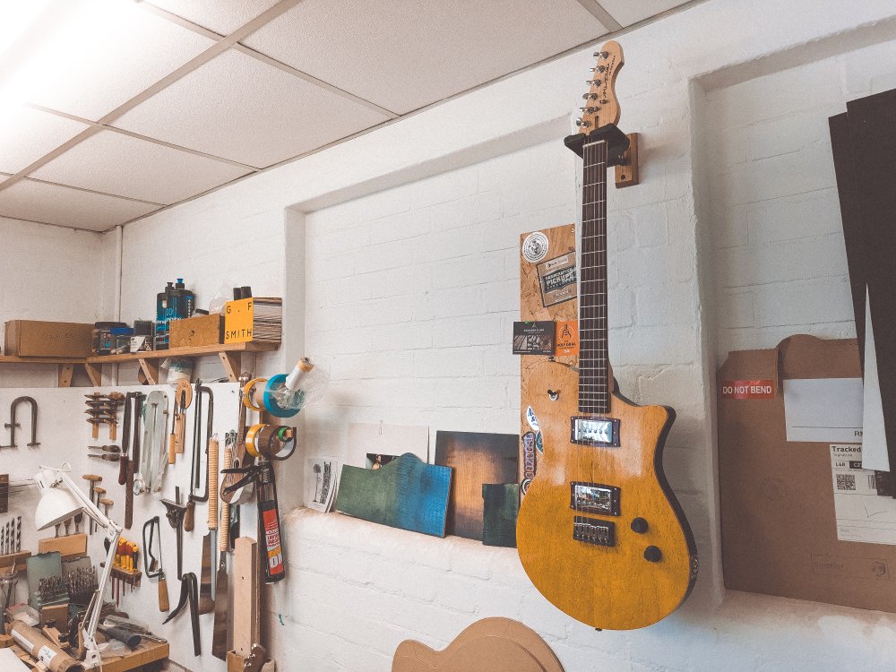 A photo of a yellow stained electric guitar hung on the wall in a workshop. Off to the left are a series of tools hung on the wall that make it look like quite a professional workshop. To the right of the guitar you can see a large cardboard envelope where the address label is mysteriously blank despite it having been franked indicating it has been throug the postal service.