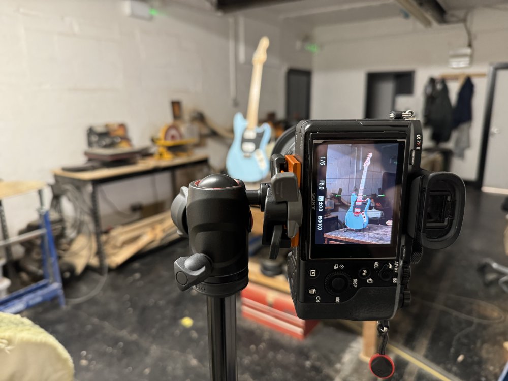 A photo of a mirrorless Sony camera on a tripod in a workshop. On the rear screen of the camera you can see in focus a guitar on a stand on a workbench, and behind the camera you can make out a blurred version of the same scene.