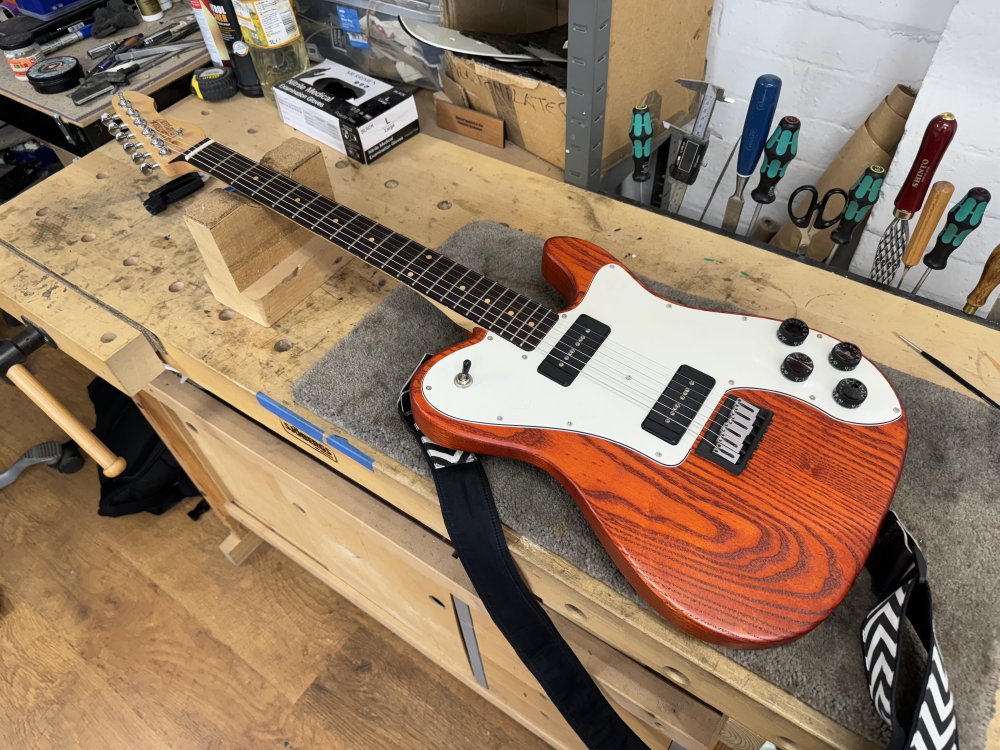 A photo of an orange telecaster-deluxe style of electric guitar sits on a workbench. The guitar has a white pick-guard with black pickups and controls, and the neck is maple with a dark wenge fretboard.