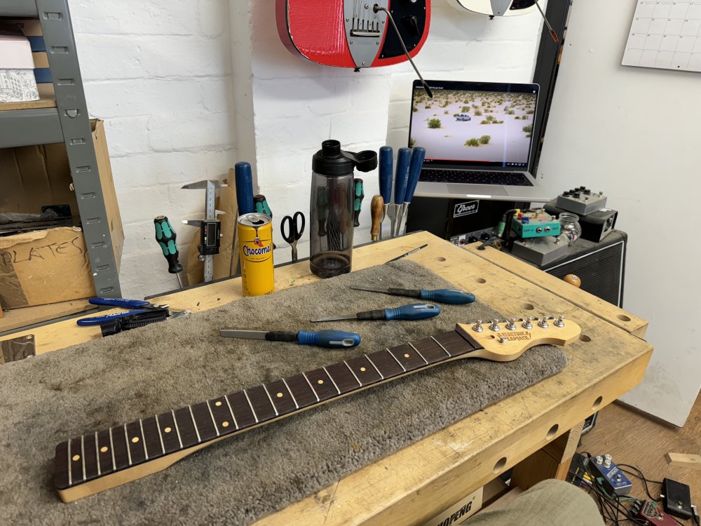 A photo of a guitar neck on a workbench surrounded by files. Behind the workbench, perched on a guitar amp, is a laptop playing a video of a car in the desert.