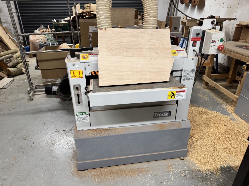 A photo of a large thicknessing machine in a workshop, with the now cleaned body blank sat on the feeder. To the right of the machine on the floor is a large pile of wood chips.