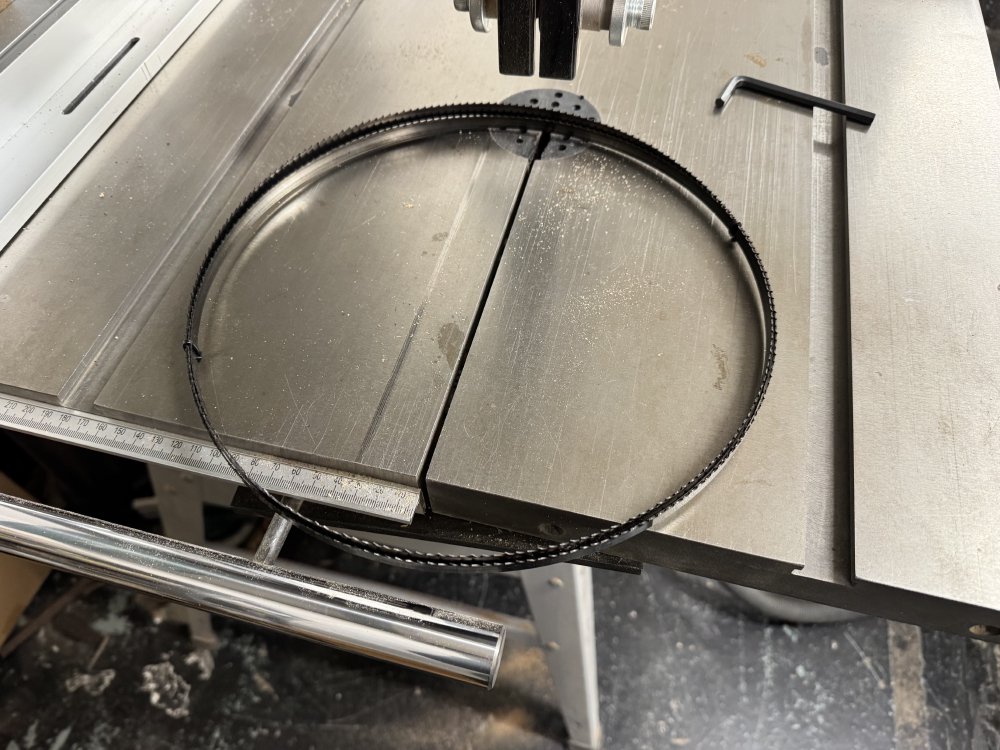 A photo of a coiled up band-saw blade sat on the table of a band-saw that currently has no blade in it. The blade is relatively thin (12mm).