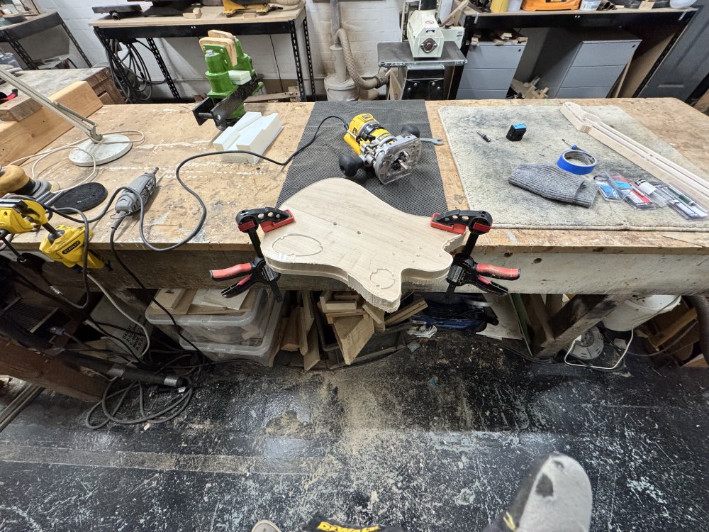 A photo of the roughed out body with template on clamped to a workbench. You can see in areas that the body has been made flush with the template, but only in short, disjoint sections, and those sections only go half the depth  of the body thickness. Behind the body on the workbench sits a Dewalt router.