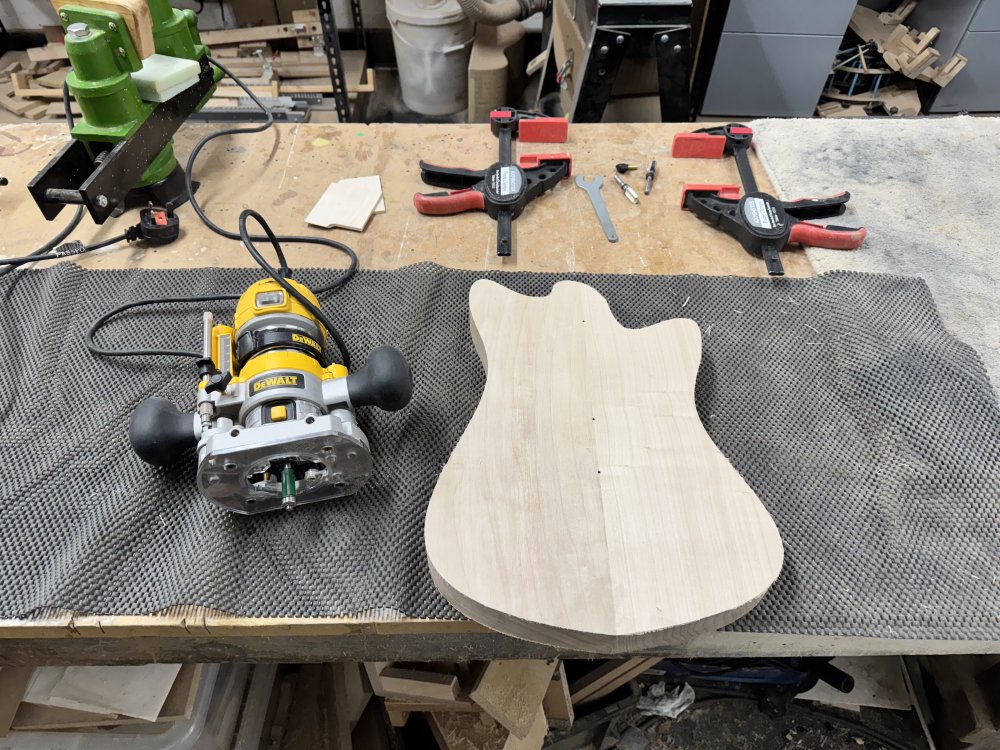 A photo of the body blank now cut down to a neat profile of a guitar body, sat on the workbench next to some clamps and a router. Notably absent from the body are any other features you'd expect, like the neck pocket, pickup cavities, etc. It is literally just cut to the profile currently.