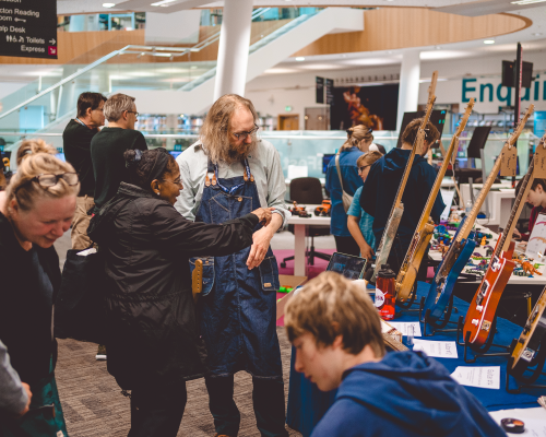 A photo of a busy library of people, with various other makers and their tables in the background, and my table with 5 guitars stood on it in the foreground. I'm stood chatting to a lady who is gesturing to one of the instruments.