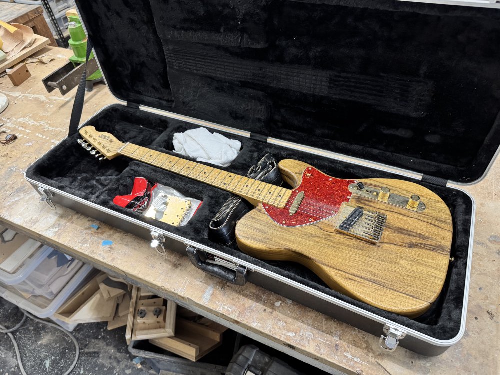 A photo of a telecaster-style electric guitar sat in a guitar case on a workbench in a workshop. The guitar has a natural wood finish on the body, which is made from figured korina, a maple neck, and has gold metal parts.