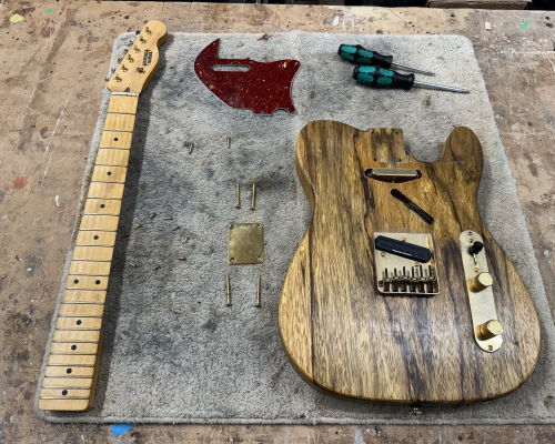 A photo of a telecaster-style electric guitar, disassembled into parts on the workbench: the neck to one side, the body to the other, the pick-guard in the middle and screws and screwdrivers around it.