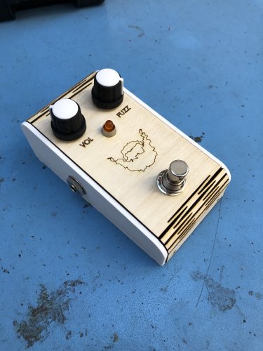 A guitar effects pedal sits on a workbench. It has two control knobs, labelled VOL and FUZZ, a footswitch, and a picture of fuzzy beard outlined on it. The lower case is 3D printed white ABS, the top surface us laser etched plywood with a living hinge letting it wrap around the top and bottom edges.