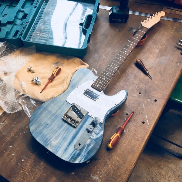 A denim blue t-style guitar sits on the workbench, with a pearloid white pickguard sporting a chrome-capped humbucker. No strings or controls yet added, this is an in-progress shot.