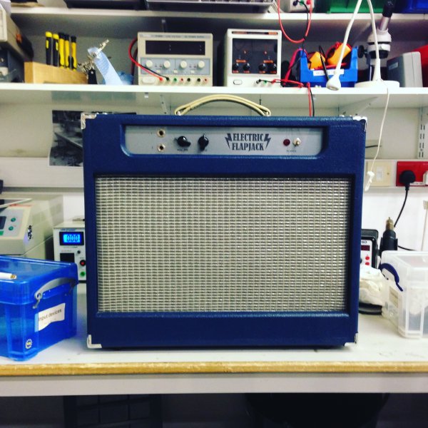 A blue all-in-one amp sits on a workbench. The main front face is taken up with silver grill fabric covering the speaker, and there is a silvery control panel with all the controls at the top.